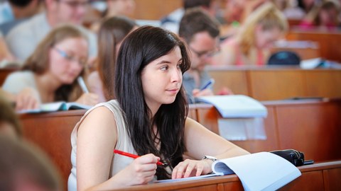 The picture shows a lecture hall with students. In the foreground a woman with dark hair.