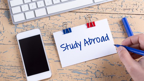 Das Foto zeigt einen Tisch. Darauf liegt eine Computertastatur, ein Handy und ein Notizzettel mit der Aufschrift "Study Abroad".