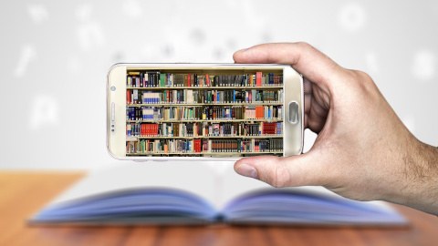 Auf einem Smartphone sieht man ein volles Bücherregal. Hinter dem Smartphone ist ein aufgeschlagenes Buch zu erkennen.