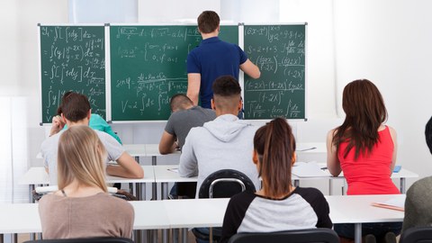 Das Foto zeigt einen Seminarraum. An den Tischen sitzen Studierende. An der Tafel löst der Dozent komplizierte mathematische Gleichungen.
