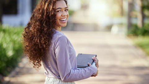 Photo of laughing student in focus standing in nature holding textbooks in hands.