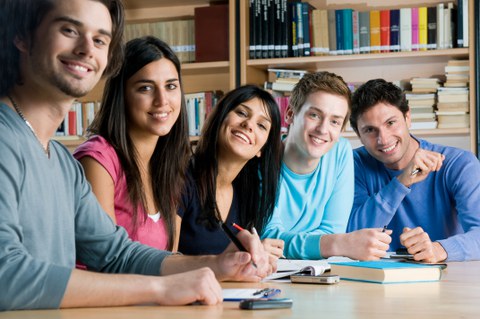 The photo shows several students sitting together at a table and smiling at the camera. In the background are bookshelves.