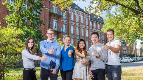 Six students stand in front of the Beyer Building of the TU Dresden and point with their thumbs up.