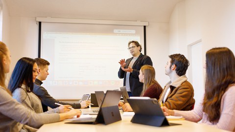 Ein Professor steht in einem Seminarraum und spricht zu den Studierenden, die vor ihm sitzen.