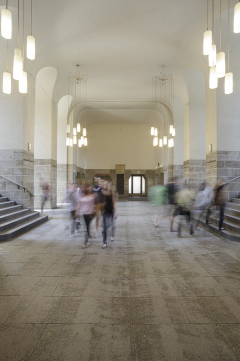 Zu sehen ist das Foyer des Georg Schumann-Baus. Es laufen Menschen durch das Foyer.   