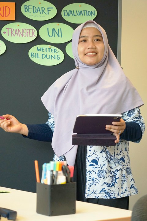 Foto einer Frau mit Kopftuch, die in einem Seminarraum mit Tablet und Stift in den Händen auf eine Tafel zeigt. 