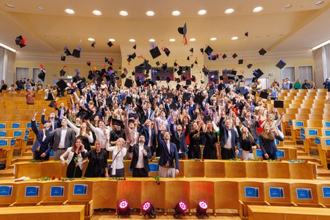 Es sind Absolventen in einem Hörsaal zu sehen, die Hüte in die Luft werfen