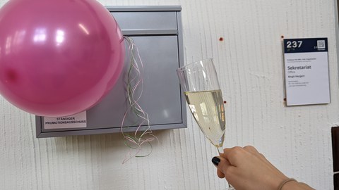 Es ist Briefkasten zu sehen, ein Luftballon und ein Glas Sekt