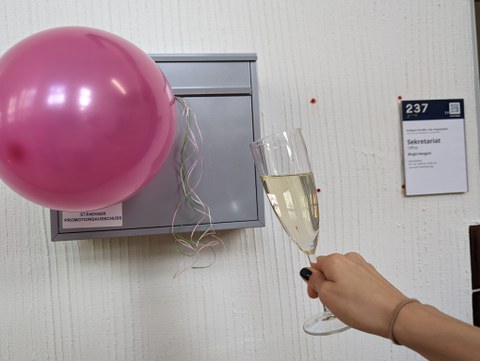 Es ist Briefkasten zu sehen, ein Luftballon und ein Glas Sekt