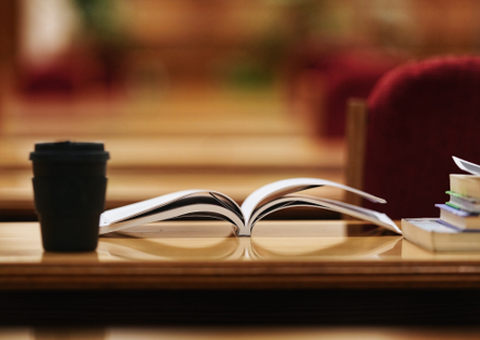 Es ist ein Tisch zu sehen, auf dem ein Kaffeebecher und ein Buch und ein Bücherstapel liegen