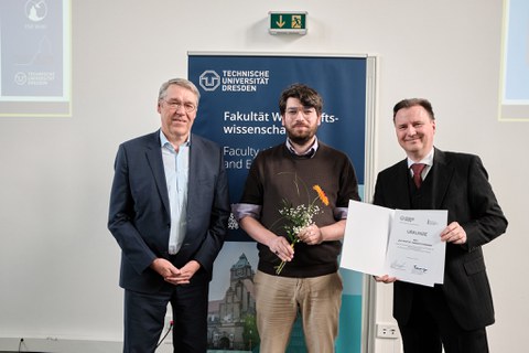 Es sind drei Männer bei einer Preisverleihung zu sehen. Der jüngere Mann in der Mitte hält eine Blume in der Hand. Er bekommt eine Urkunde überreicht  