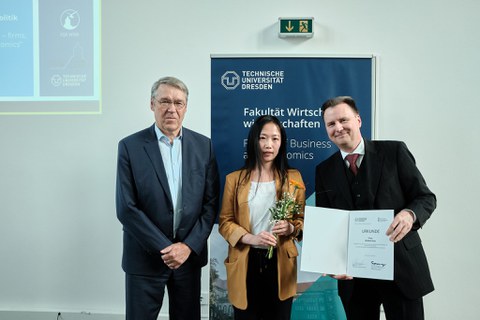 Es sind eine Frau und zwei Männer bei einer Preisverleihung zu sehen. Die Frtau hält eine Blume in der Hand. Sie bekomm eine Urkunde überreicht