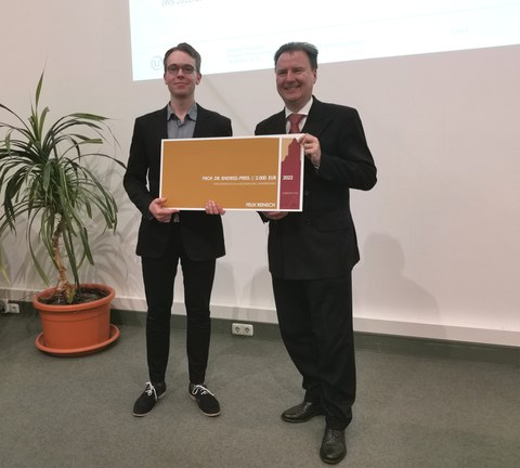 Es ist der Dekan zu sehen und ein Student. Beide halten einen großen farbigen Scheck in der Hand und lachen. Es handelt sich um eine Preisverleihung   
