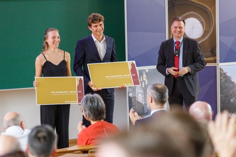 Zu sehen sind zwei Studierende und der Dekan. Die beiden Studierenden halten große Schecks in der Hand und lachen. Es handelt sich um eine Preisverleihung  