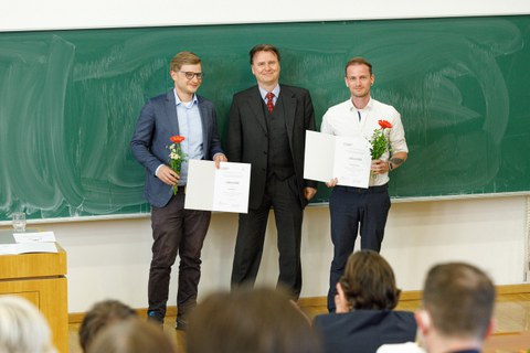 Es sind drei Männer zu sehen, die in einem Hörsaal vor einer Tafel stehen. In der Mitte steht der Dekan neben zwei Preisträgern, die Urkunden in den Händen halten