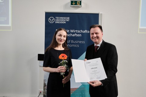 Es sind eine junge Frau und ein Mann zu sehen, die vor einem Rollup stehen. Es handelt sich um eine Preisverleihung. Sie halten eine Blume und eine Urkunde in der Hand  