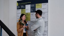 Zu sehen sind zwei junge Menschen, die sich vor einer Infotafel unterhalten 