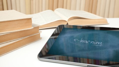 Das Foto zeigt ein Tablett, auf dem das Wort "e-learning" abgebildet ist. Das Tablett liegt gemeinsam mit einigen Büchern auf einem Tisch.