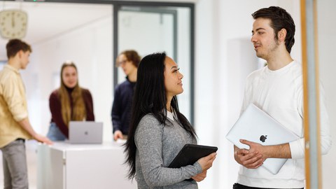 Im Vordergrund unterhalten sich eine junge Frau und ein junger Mann. Im Hintergrund reden drei Studierende miteinander im Gang.