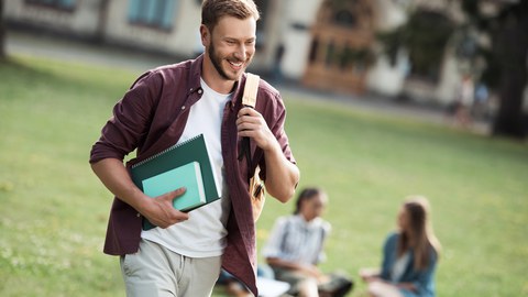 Foto eines Studenten, der Bücher in der Hand hält und lächelnd an zwei auf der Wiese sitzenden Studentinnen vorbeiläuft.