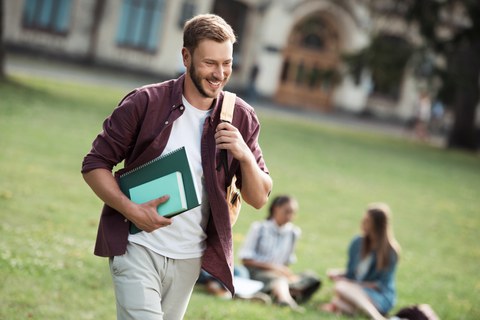 Foto eines Studenten, der Bücher in der Hand hält und lächelnd an zwei auf der Wiese sitzenden Studentinnen vorbeiläuft.