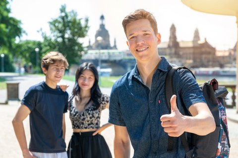 Das Bild zeigt drei Studenten, die an der Elbe mit Blick auf Dresden stehen und in die Kamera lächeln. Einer davon macht eine Daumen-hoch Geste