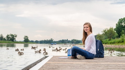 oto: Eine Studentin sitzt auf einem Bootssteg an der Elbe und Enten tummeln sich auf dem Wasser.