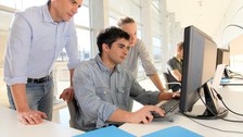 Das Foto zeigt zwei Studierende vor einem Computer. Der Junge sitzt auf dem Stuhl und tippt auf der Tastatur. Der Dozent steht daneben und schaut ebenfalls auf den Bildschirm.