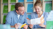 Das Foto zeigt zwei Personen an einem Tisch. Sie halten Informationsflyer zu verschiedenen Studiengängen in den Händen, schauen jedoch gemeinsam in einen der Flyer. Im Hintergrund erkennt man ein Regal mit weiterem Informationsmaterial zu den verschiedenen Studiengängen.