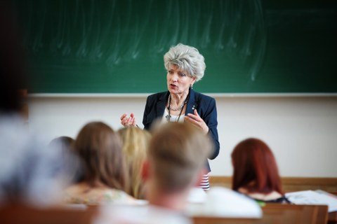 Das Foto zeigt eine Dozentin vor ihrem Kurs. Sie steht im Fokus des Bildes und spricht zu den Studierenden. Im Hintergrund erkennt man eine grüne Kreidetafel