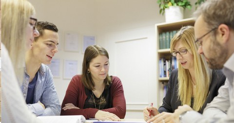 Studierende und Dozierende sitzen an einem Tisch und besprechen ein Thema.