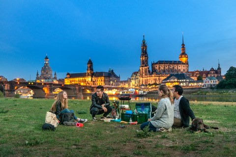 Foto: Vier Studenten sitzen mit einem Hund am Abend an den Elbwiesen von Dresden und grillen.