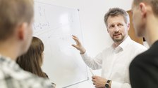 Das Foto zeigt eine Tafel, an der Formel und Grafiken geschrieben stehen. Vor der Tafel steht der Dozent, der drei Studierenden etwas erklärt.