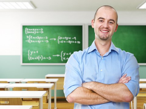 Das Foto zeigt einen Lehrer in seinem leeren Klassenraum. Er hat die Arme vor dem Körper verschränkt und lächelt in die Kamera.