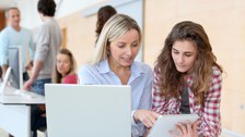 Im Fokus des Fotos stehen zwei Frauen. Während die eine Frau vor einem Laptop sitzt, hat die andere ein Tablet in der Hand. Die Frau mit dem Laptop zeigt der anderen etwas auf ihrem Tablet.