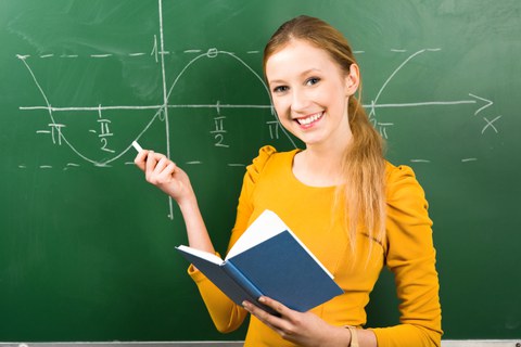 Das Foto zeigt eine junge Frau mit einem Buch in der Hand, die vor einer Kreidetafel steht. An die Tafel ist eine Sinuskurve gezeichnet.
