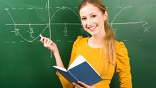 Das Foto zeigt eine junge Frau mit einem Buch in der Hand, die vor einer Kreidetafel steht. An die Tafel ist eine Sinuskurve gezeichnet.