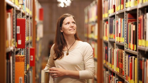 Das Foto zeigt eine Studentin in der Bibliothek. Sie steht zwischen den Bücherregalen, hält einige Bücher unter ihrem Arm und blickt in eine Regalreihe