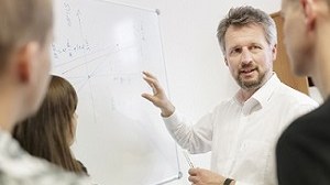 Zu sehen ist ein Professor, der drei Studierenden etwas an einem Whiteboard erklärt