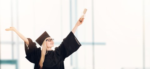 Auf dem Foto ist eine Frau zu sehen, die einen Doktorhut trägt und lächelnd die Hände in die Luft hält. In der linken Hand hält sie ein Diplom.