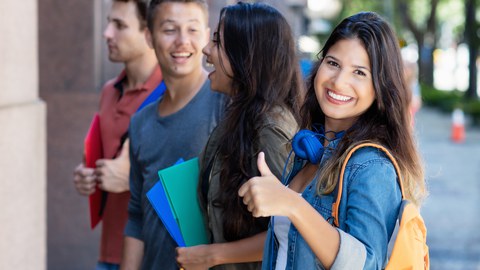 Vier junge, fröhliche Menschen stehen perspektivisch in einer Reihe. Die Studentin an erster Stelle lächelt in die Kamera und hält den gestreckten Daumen hoch