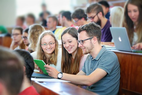 Das Foto zeigt drei Studierende in einem vollen Hörsaal. Sie sitzen nebeneinander und schauen gemeinsam in ein Notebook