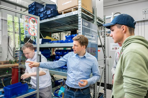 Foto eines Dozenten, der auf ein Gerät hinter einer Plexiglasglasscheibe zeigt. Er wird von drei seiner Studenten dabei beobachtet.