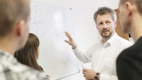Photo of Prof. Kemnitz in front of a whiteboard explaining something to three students.