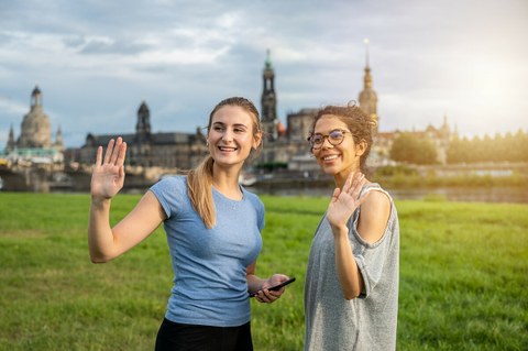 Foto zeigt zwei lächelnde und winkende Studentinnen an der Elbe.