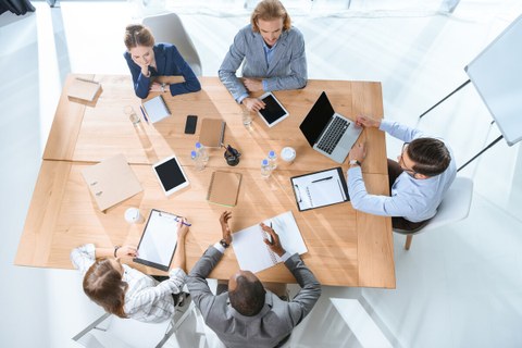 Bild von fünf Personen, die an einem großen Tisch sitzen und zusammen arbeiten aus der Vogelperspektive.