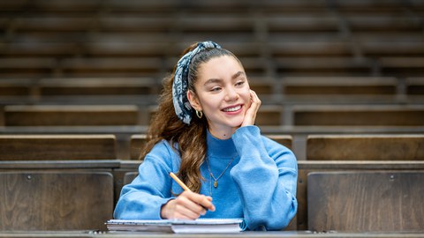 Foto einer Studierenden, die glücklich lächelnd im Hörsaal sitzt.