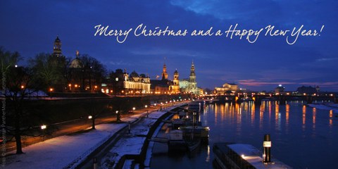 Foto der Dresden Silhouette mit Blick auf Elbe in Richtung Semperoper in weihnachtlich beleuchteter Abendstimmung mit spiegelnden Lichtern im Fluss und dem Schriftzug in Schreibschrift: "Merry Christmas and a Happy New Year!" 