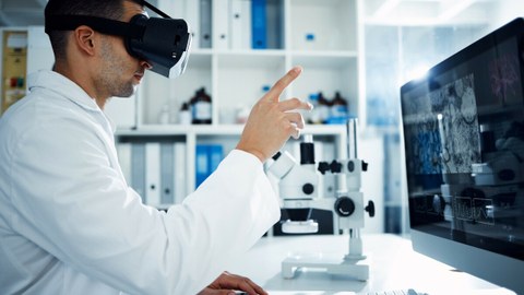 Foto eines Mannes im weißen Laborkittel, der vor einem Bildschirm sitzt und durch eine VR-Brille sieht.