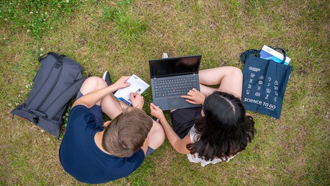 Bild von zwei Studenten, die auf der Wiese zusammen am Laptop arbeiten.